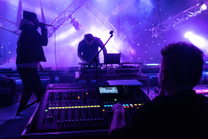Back of a student sat at a mixing desk with sliders and coloured lights. In the background is a DJ with decks and a laptop, with smoke and purple and blue stage lighting